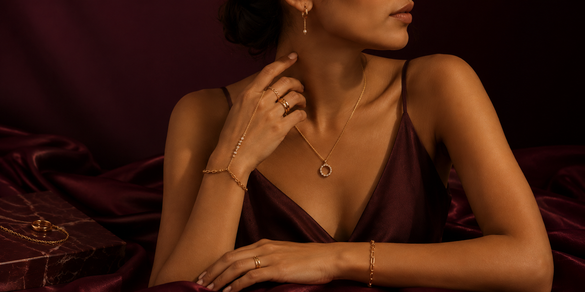 A woman in a dark burgundy satin dress models a delicate gold necklace with a circular pendant, matching gold bracelets, and rings, set against a dark burgundy satin background.